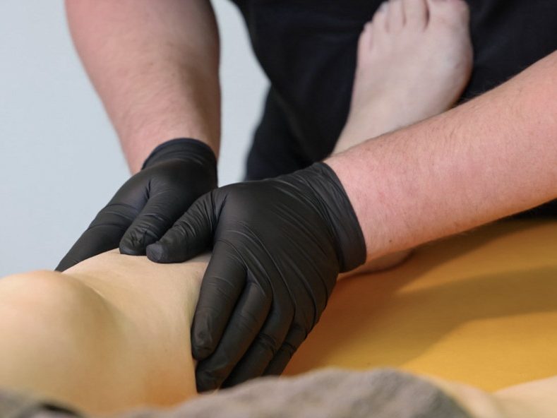 Eine Person mit schwarzen Handschuhen führt eine Massage an einem Bein durch.